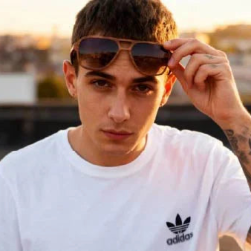 A man wearing a white Adidas t-shirt and aviator sunglasses, looking directly at the camera with a blurred outdoor background.
