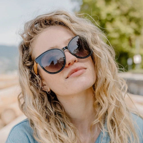 A close-up lifestyle shot of a woman with wavy blonde hair wearing Santorini sunglasses and a light blue denim shirt, set against a soft-focus outdoor background.