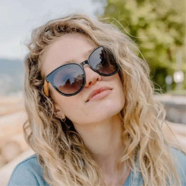 A woman with long wavy blonde hair wearing Wildwood Santorini sunglasses and a light blue denim shirt