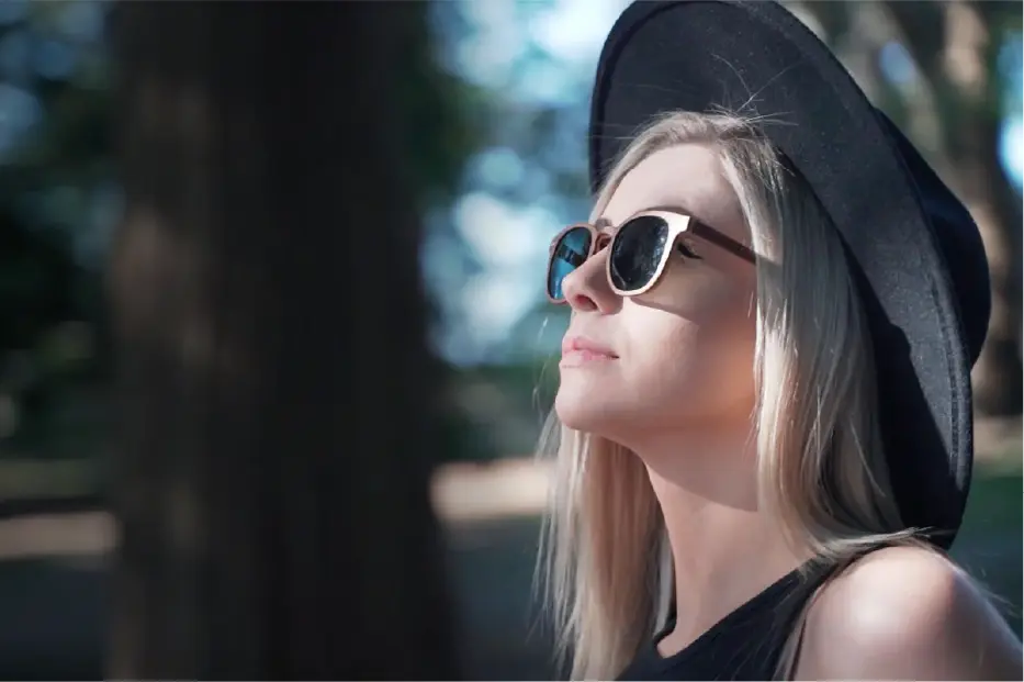 A woman in a black wide-brim hat and black sunglasses with white frames, shown in profile outdoors against a blurred background of trees.